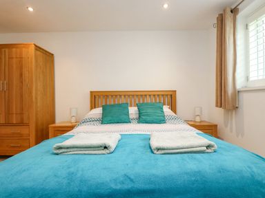 A bedroom with a wooden bed frame two teal pillows folded towels on a blue blanket wooden wardrobe and bedside tables with lamps at Rock Pool in Downderry
