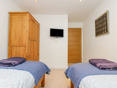 A bedroom with two single beds, wooden wardrobe, wall-mounted TV, wooden door, and wall artwork at Rock Pool in Downderry