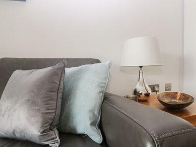 A grey sofa with two cushions next to a wooden side table with a lamp and decorative bowls at Rock Pool in Downderry