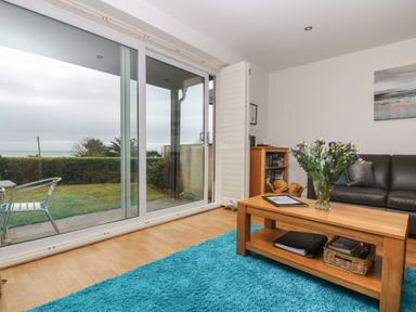 A living room with a wooden coffee table and vase of flowers next to a black sofa and large sliding glass doors at Rock Pool in Downderry