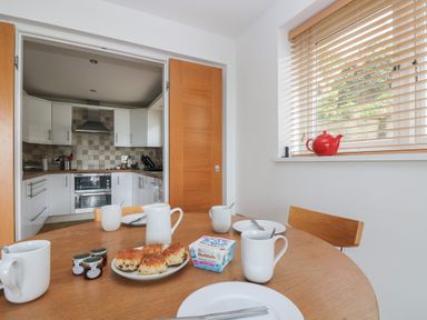 A kitchen with white cabinets and tiled backsplash seen through open wooden doors from a dining area with a table set with cups plates scones and jam at Rock Pool in Downderry