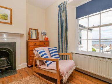 A room with a wooden rocking chair with cushions and a blanket next to a wooden chest of drawers and a window overlooking rooftops and water at 1 Dormer Villas in Portscatho