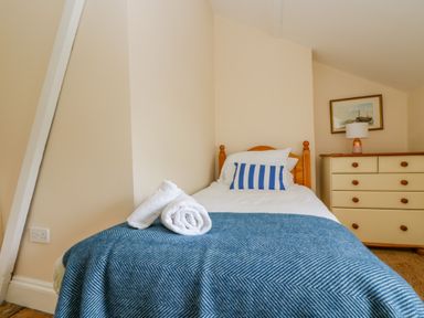 A bedroom with a single bed with rolled towels and a striped pillow next to a dresser with a lamp and framed picture at 1 Dormer Villas in Portscatho