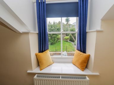 A window seat with two yellow cushions blue curtains and a view of a garden at 1 Dormer Villas in Portscatho