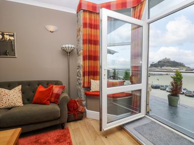 A living room with a sofa and cushions next to an open patio door showing a view of a castle and parked cars at Mount View in Marazion