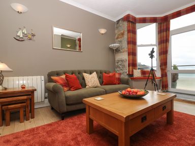 A living room with a green sofa and red cushions a wooden coffee table with a bowl of fruit a side table with nested stools and a lamp a wall mirror and wall art a telescope by the window and a red and orange curtain at Mount View in Marazion