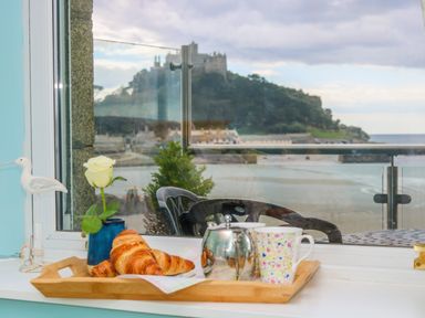 A window sill with a tray holding croissants a teapot and a floral mug a white rose in a blue vase and a bird figurine by a window overlooking a castle hill and sea at Mount View in Marazion