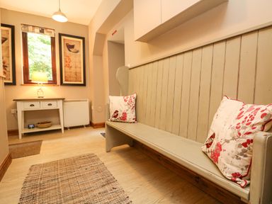A hallway with a wooden bench with floral cushions a small table with a lamp two framed pictures on the wall and a woven rug on the floor at Chenalls Cottage in Helston