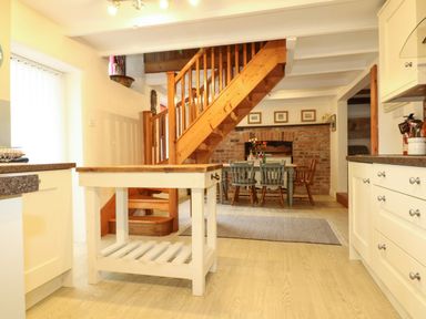 A kitchen with white cabinets a wooden island and a dining area with wooden chairs and a brick wall at Chenalls Cottage in Helston