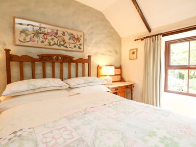 A bedroom with a wooden bed frame bedside table with lamp and window with curtains at Chenalls Cottage in Helston