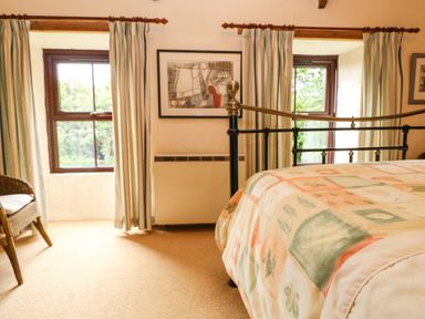 A bedroom with a metal bed frame patterned bedspread two windows with striped curtains a wicker chair and framed artwork at Chenalls Cottage in Helston