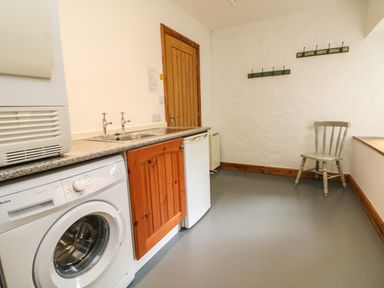 A utility room with a washing machine sink wooden cabinet small fridge chair and wall hooks at Chenalls Cottage in Helston
