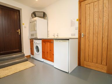 A utility room with a washing machine, dryer, small refrigerator, wooden cabinets and two doors at Chenalls Cottage in Helston