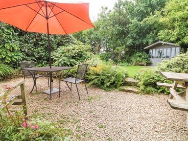 An outdoor patio with a table and two chairs under an orange umbrella next to a wooden bench and a wooden picnic table in a garden with plants and a gray garden shed in the background at Chenalls Cottage in Helston