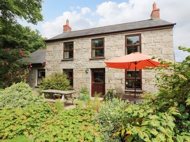 A stone house with wooden framed windows a wooden door a picnic table and an outdoor table with an orange umbrella surrounded by plants and bushes at Chenalls Cottage in Helston