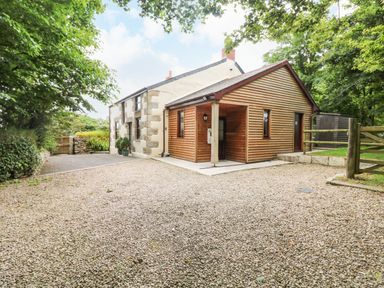 A house with wooden and stone exterior surrounded by trees and gravel driveway at Chenalls Cottage in Helston