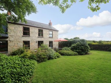 A stone house with multiple windows a garden with bushes green grass and a patio table with an orange umbrella at Chenalls Cottage in Helston