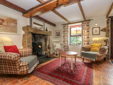 A living room with a stone fireplace two armchairs a rocking chair a red rug and a window with patterned curtains at Chenalls Cottage in Helston