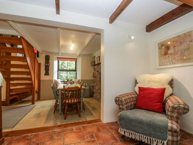A dining area with wooden chairs and table near a wooden staircase and a plaid armchair with cushions at Chenalls Cottage in Helston
