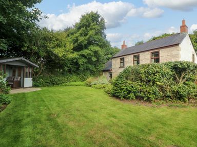 A garden with grass lawn bushes a small building and a stone house at Chenalls Cottage in Helston