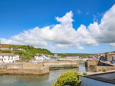 A coastal village with stone piers extending into the water surrounded by houses and green hills under a blue sky at Chenalls Cottage in Helston