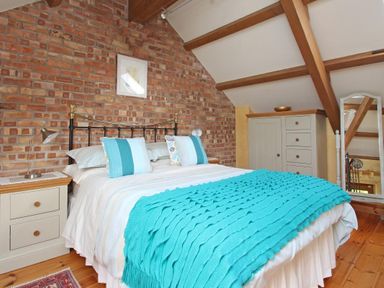 A bedroom with a bed covered with a turquoise blanket and pillows against a brick wall with wooden furniture and a standing mirror at Summer House in Helston