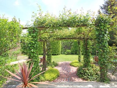 A garden with a brick circular path and wooden pergola covered with greenery and plants at Summer House in Helston