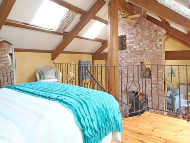 A bedroom loft with wooden beams a brick chimney a ceiling fan a wicker chair and a bed with a blue throw at Summer House in Helston