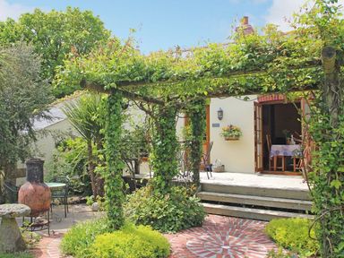 A garden patio with a wooden pergola covered in vines a brick circular patterned floor chairs a table and an open door leading inside at Summer House in Helston