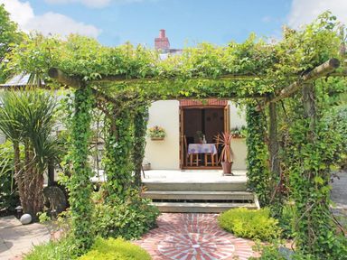 A garden with a wooden pergola covered in vines leading to a patio with a table and chairs at Summer House in Helston