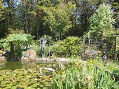 A garden pond with lily pads and statues next to a bench and trees at Summer House in Helston