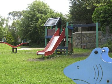 A playground with a slide, swing, seesaw, and shark-shaped play equipment surrounded by grass and trees at White Duck Cottage in Hayle
