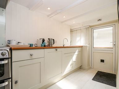 A kitchen with cream cabinets a wooden countertop a kettle coffee machine cups a sink a door with a cat flap and a window with blinds at Mallards in Pentewan