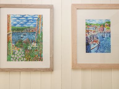 Two framed paintings of harbor scenes with boats and buildings on a cream paneled wall at Mallards in Pentewan