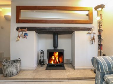 A fireplace with a burning wood stove and fire tools next to a wicker basket and a striped armchair at Mallards in Pentewan