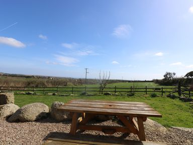 A wooden picnic table on gravel with a green field and a wooden fence under a blue sky at Boar's House in Penzance