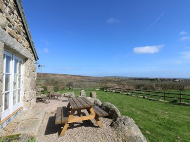 A stone house with a wooden picnic table and chairs outside overlooking a grassy field and distant hills at Boar's House in Penzance