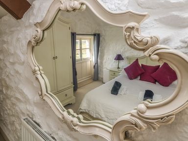 A bedroom with a bed red pillows a wardrobe bedside table and a lamp seen in a decorative mirror at Boar's House in Penzance