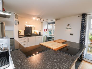 A kitchen with black countertops, two wooden cutting boards, white cabinets, a toaster, and a window with blue patterned curtains at Ebenezer Cottage in Marazion