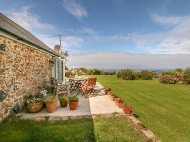 A stone house with a patio table and chairs and flower pots on a lawn with bushes and ocean view at Back Of Beyond in St Ives