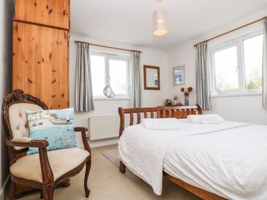 A bedroom with a wooden bed, bench, two windows with curtains, a wooden chair with a pillow, and a wooden wardrobe at Angarrack Farmhouse in Hayle