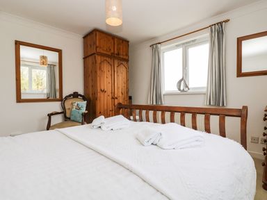A bedroom with a wooden bed, white linens, a wooden wardrobe, an armchair with a cushion, two mirrors on the wall, and a window with curtains at Angarrack Farmhouse in Hayle