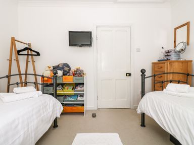 A bedroom with two single beds with white bedding a wooden chest of drawers a small shelf with toys and books a wall-mounted television and a wooden clothes rack at Angarrack Farmhouse Hayle