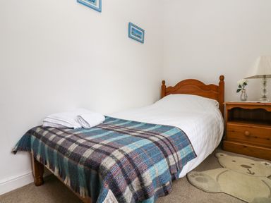 A single bed with a blue plaid blanket and white towels on top next to a wooden nightstand with a lamp and a flower in a vase at Angarrack Farmhouse in Hayle