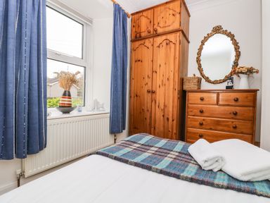 A bedroom with wooden wardrobe and chest of drawers blue curtains and a bed with a plaid blanket and white towels at Angarrack Farmhouse in Hayle