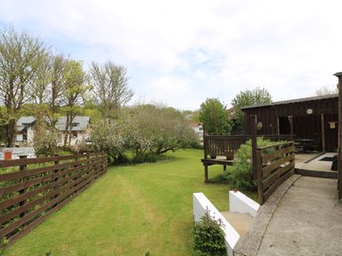 A garden with green lawn and trees next to a wooden fence and a wooden deck with seating at Angarrack Farmhouse in Hayle