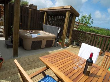 An outdoor deck area with a hot tub under a wooden pergola a wooden table with wine bottle and glasses and wooden chairs at Godrevy Cottage in Hayle