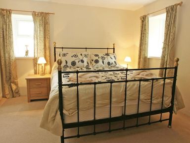 A bedroom with a metal bed frame a floral bedspread bedside tables with lamps and windows with curtains at Godrevy Cottage in Hayle