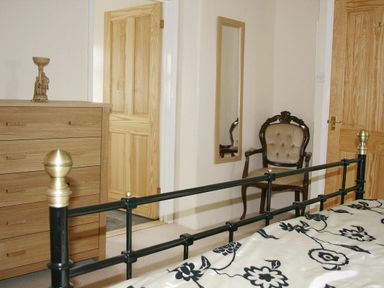 A bedroom with a black metal bed frame a wooden chest of drawers an antique upholstered chair and a wall mirror at Godrevy Cottage in Hayle