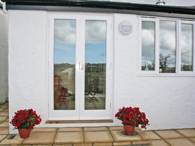 White double glass doors with two potted red flowering plants on a tiled patio at Godrevy Cottage in Hayle